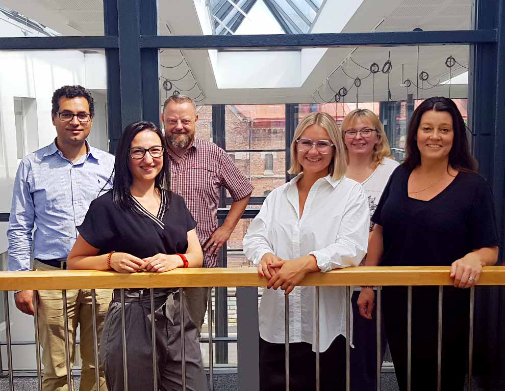 6 Plasman EU Headquarters employees standing behind wood railing in front of large glass windows at Headquarters in Gothenburg, Sweden
