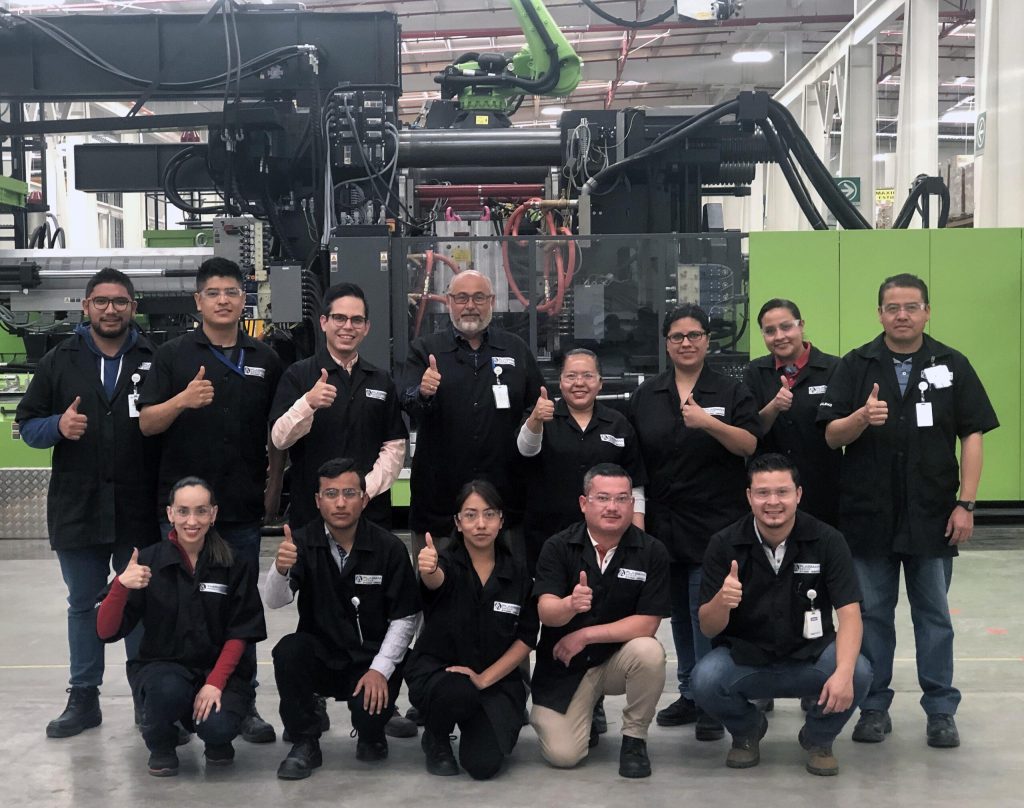 Group of employees from Plasman Queretaro, Mexico posing in front of a large green molding machine holding up their thumbs in a #1 gesture