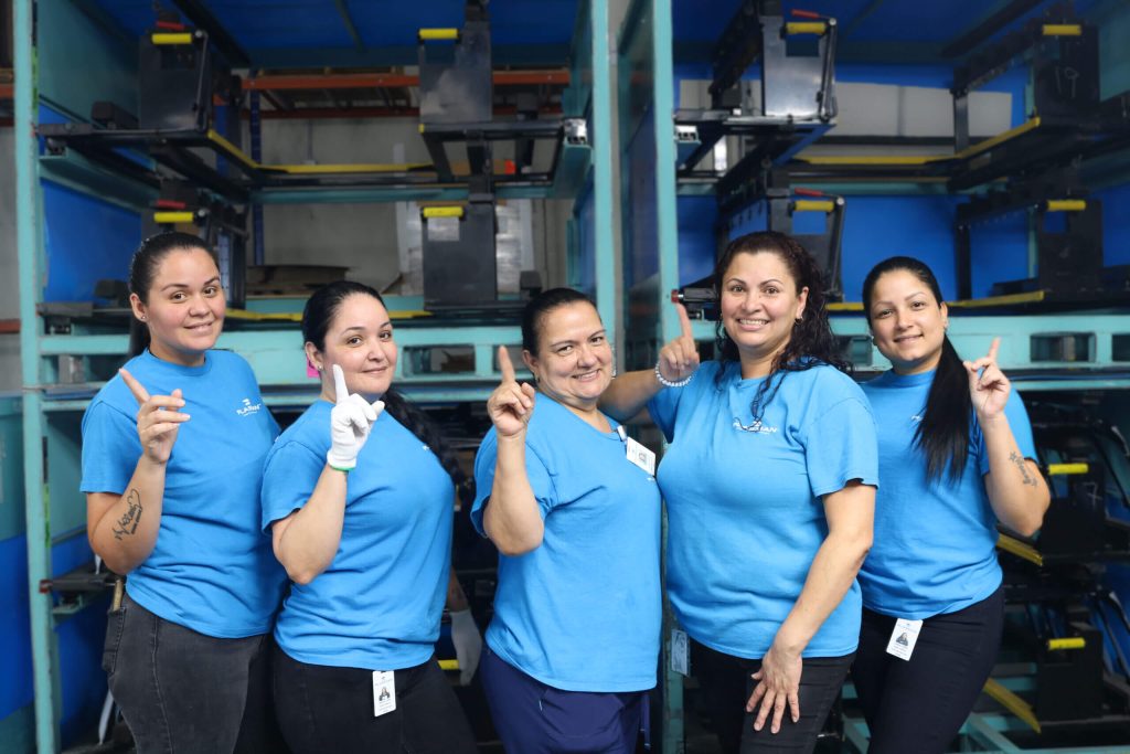 Group of five women wearing cyan Plasman shirts holding up the number 1 finger and smiling in front of storage bins for exterior trim parts