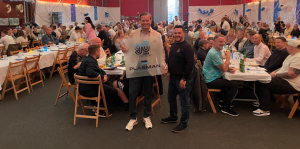 Picture of Patirk plant manager of Simrishamn holding a 90th Anniersary plaque with employees sitting at tables behind him