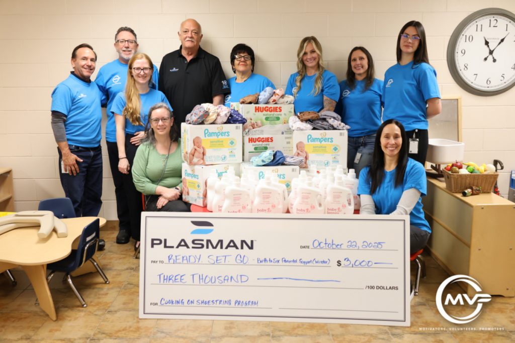 Group of people gathered around a table full of baby diapers and wipes with a $3,000 big cheque in front of table