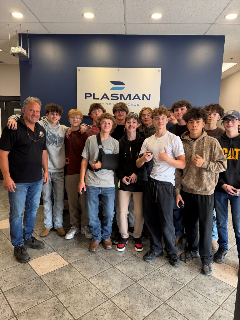 A bunch of students gathered in the lobby of a Plasman facility standing in front of a Plasman sign